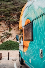 A colorful camper van with vibrant designs painted on its side is parked by a cliffside with lush greenery. The van is predominantly turquoise with an orange and white design near the roof and window. The side mirror is visible, and the background features a sandy area and vegetation.
