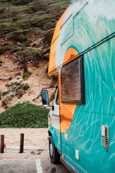 A colorful camper van with vibrant designs painted on its side is parked by a cliffside with lush greenery. The van is predominantly turquoise with an orange and white design near the roof and window. The side mirror is visible, and the background features a sandy area and vegetation.