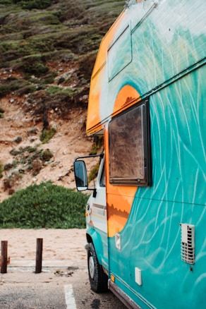 A colorful camper van with vibrant designs painted on its side is parked by a cliffside with lush greenery. The van is predominantly turquoise with an orange and white design near the roof and window. The side mirror is visible, and the background features a sandy area and vegetation.