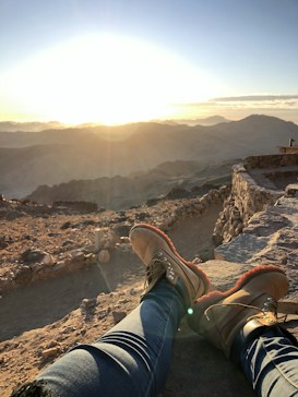 A scenic view of a mountainous landscape at sunrise or sunset with warm sunlight illuminating the horizon. The foreground features a person wearing jeans and hiking boots resting on a rocky surface, while stone structures or pathways are visible on the right side.