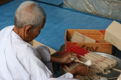 An elderly person in white clothing is sorting and bundling incense sticks on a blue surface. There are open cardboard boxes filled with incense sticks nearby, and the person is using a small tool to organize them.