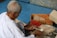 An elderly person in white clothing is sorting and bundling incense sticks on a blue surface. There are open cardboard boxes filled with incense sticks nearby, and the person is using a small tool to organize them.