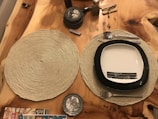 A wooden table with two woven placemats, one of which holds a square plate with a black border and subtle line designs in the center. On the placemat, there is also a set of utensils, including a fork, spoon, and knife. Nearby, several coasters are stacked, a small bottle is placed above them, and the corner of a magazine is visible.
