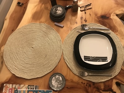A wooden table with two woven placemats, one of which holds a square plate with a black border and subtle line designs in the center. On the placemat, there is also a set of utensils, including a fork, spoon, and knife. Nearby, several coasters are stacked, a small bottle is placed above them, and the corner of a magazine is visible.