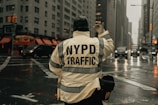 A person wearing a jacket with 'NYPD Traffic' written on the back is directing traffic at an intersection in a city. The scene is urban with tall buildings, and there is a visible wet street, suggesting recent rain. Vehicles are waiting, and the traffic lights can be seen in the background.