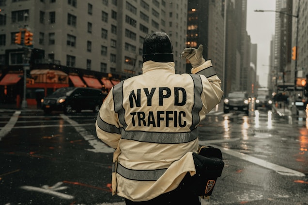 A person wearing a jacket with 'NYPD Traffic' written on the back is directing traffic at an intersection in a city. The scene is urban with tall buildings, and there is a visible wet street, suggesting recent rain. Vehicles are waiting, and the traffic lights can be seen in the background.