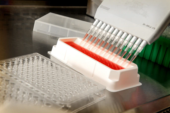 Multichannel pipette drawing red liquid from a reagent reservoir next to a 96-well microplate for hi