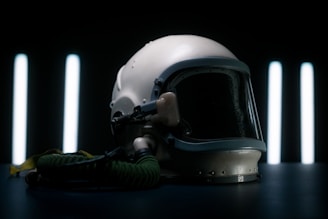 A vintage aviation helmet with a dark visor and attached oxygen hose is placed on a dark surface. The background features vertical, glowing light strips, creating a dramatic and futuristic ambiance.