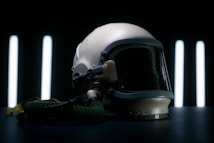 A vintage aviation helmet with a dark visor and attached oxygen hose is placed on a dark surface. The background features vertical, glowing light strips, creating a dramatic and futuristic ambiance.