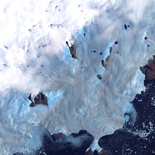 A satellite view captures a frozen landscape dominated by vast, swirling white patterns of ice and snow. Dark brown landforms scattered throughout contrast sharply with the bright icy surface. Patches of deep blue water appear sporadically, suggesting meltwater or small lakes. The bottom right corner features a dark, textured expanse, possibly indicating open water or sea ice.