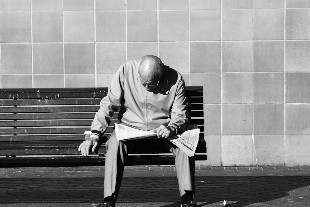 grayscale photography of man sitting on bench while reading newspaper