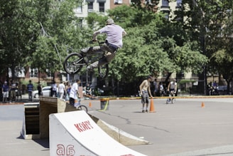 A person on a BMX bike performing a jump off a ramp in an outdoor skate park. Several people stand in the background, some watching, while others participate in activities. The scene is set on a sunny day with trees and buildings visible in the background.