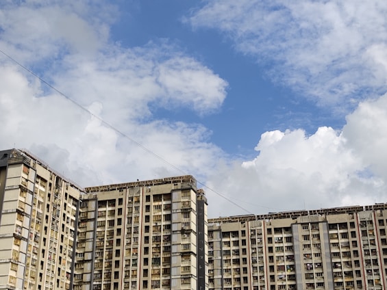 High-rise residential buildings with multiple floors and balconies stand against a bright, partly cloudy sky. The architecture is characterized by a uniform pattern of windows and visible exterior wear.