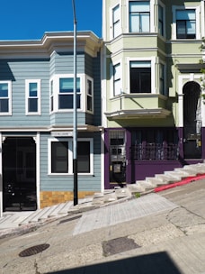 Two adjacent, multi-story residential buildings are situated on a steeply inclined street. The left building is painted in gray with white trim and has bay windows. The right building features a light green facade with a dark purple lower section and an ornate black wrought iron gate. A concrete sidewalk and street run diagonally in the foreground. A street sign is visible on the left side of the image.