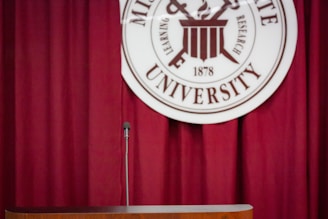 A dark red curtain serves as the backdrop for a podium with a microphone. Behind and slightly above, there is a circular university seal with text and symbols.