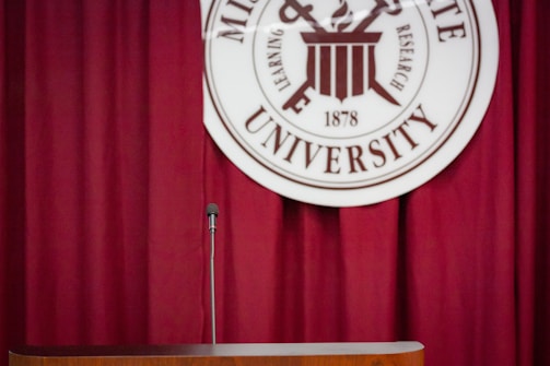 A dark red curtain serves as the backdrop for a podium with a microphone. Behind and slightly above, there is a circular university seal with text and symbols.