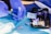 A pair of gloved hands handling a syringe and a medical testing device on a blue surface. The scene includes a digital blood glucose meter in a black pouch, test strips, and packaged medical supplies. The setting appears clinical or medical in nature, emphasizing the process of testing or preparation.
