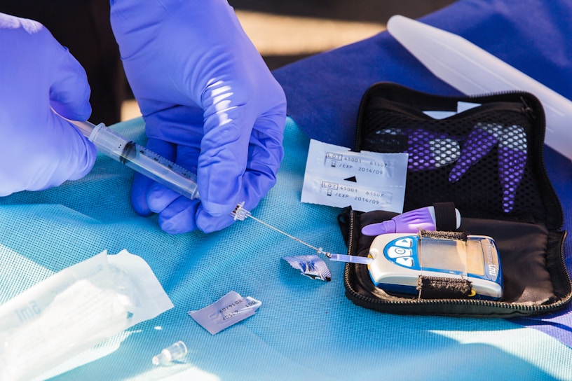 A pair of gloved hands handling a syringe and a medical testing device on a blue surface. The scene includes a digital blood glucose meter in a black pouch, test strips, and packaged medical supplies. The setting appears clinical or medical in nature, emphasizing the process of testing or preparation.