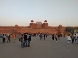 A large historic red sandstone fort with multiple domes and towers stands prominently in the background. Numerous people are walking and gathering in the open area in front of the fort, some taking photos or talking in groups. The sky is clear and the scene appears lively and bustling.