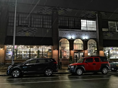 A nighttime scene in front of the Denver Central Market, featuring two parked cars on a wet street. The market's facade includes large, arched windows illuminated from inside. Light decorations are visible, creating a warm and inviting glow against the dark surroundings. Geometric patterns adorn the upper walls of the building.