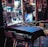 An outdoor dining area features metal chairs and tables covered with dark tablecloths. The scene includes a window adorned with various posters and a string of colorful lights, contributing to a festive atmosphere. Reflections on the glass showcase a neon-lit interior with a portrait.