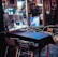 An outdoor dining area features metal chairs and tables covered with dark tablecloths. The scene includes a window adorned with various posters and a string of colorful lights, contributing to a festive atmosphere. Reflections on the glass showcase a neon-lit interior with a portrait.