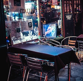 An outdoor dining area features metal chairs and tables covered with dark tablecloths. The scene includes a window adorned with various posters and a string of colorful lights, contributing to a festive atmosphere. Reflections on the glass showcase a neon-lit interior with a portrait.