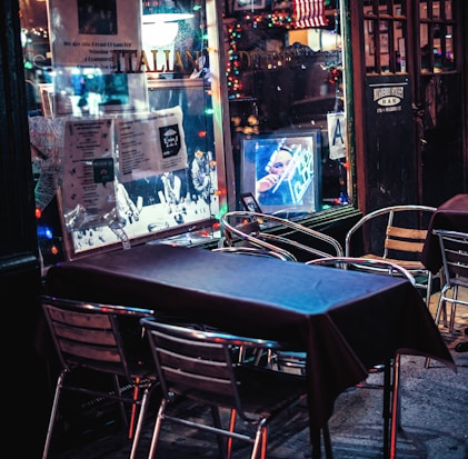 An outdoor dining area features metal chairs and tables covered with dark tablecloths. The scene includes a window adorned with various posters and a string of colorful lights, contributing to a festive atmosphere. Reflections on the glass showcase a neon-lit interior with a portrait.