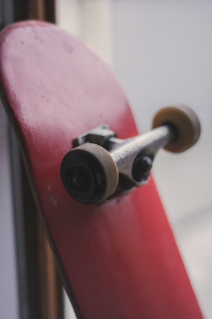 A red skateboard is resting vertically against a surface with a focus on the underside. The skateboard has two visible wheels and the metal truck assembly attached. The background is blurred, emphasizing the skateboard as the main subject.