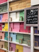 Shelves filled with colorful bars of soap, neatly arranged by color. Each bar is labeled with different scents or ingredients, and there is a chalkboard sign displaying pricing information. Some of the prominent colors include pink, yellow, green, and blue.