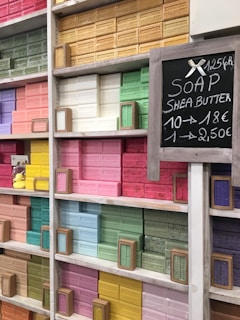Shelves filled with colorful bars of soap, neatly arranged by color. Each bar is labeled with different scents or ingredients, and there is a chalkboard sign displaying pricing information. Some of the prominent colors include pink, yellow, green, and blue.