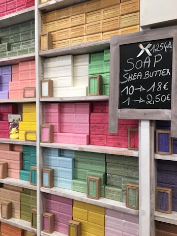 Shelves filled with colorful bars of soap, neatly arranged by color. Each bar is labeled with different scents or ingredients, and there is a chalkboard sign displaying pricing information. Some of the prominent colors include pink, yellow, green, and blue.