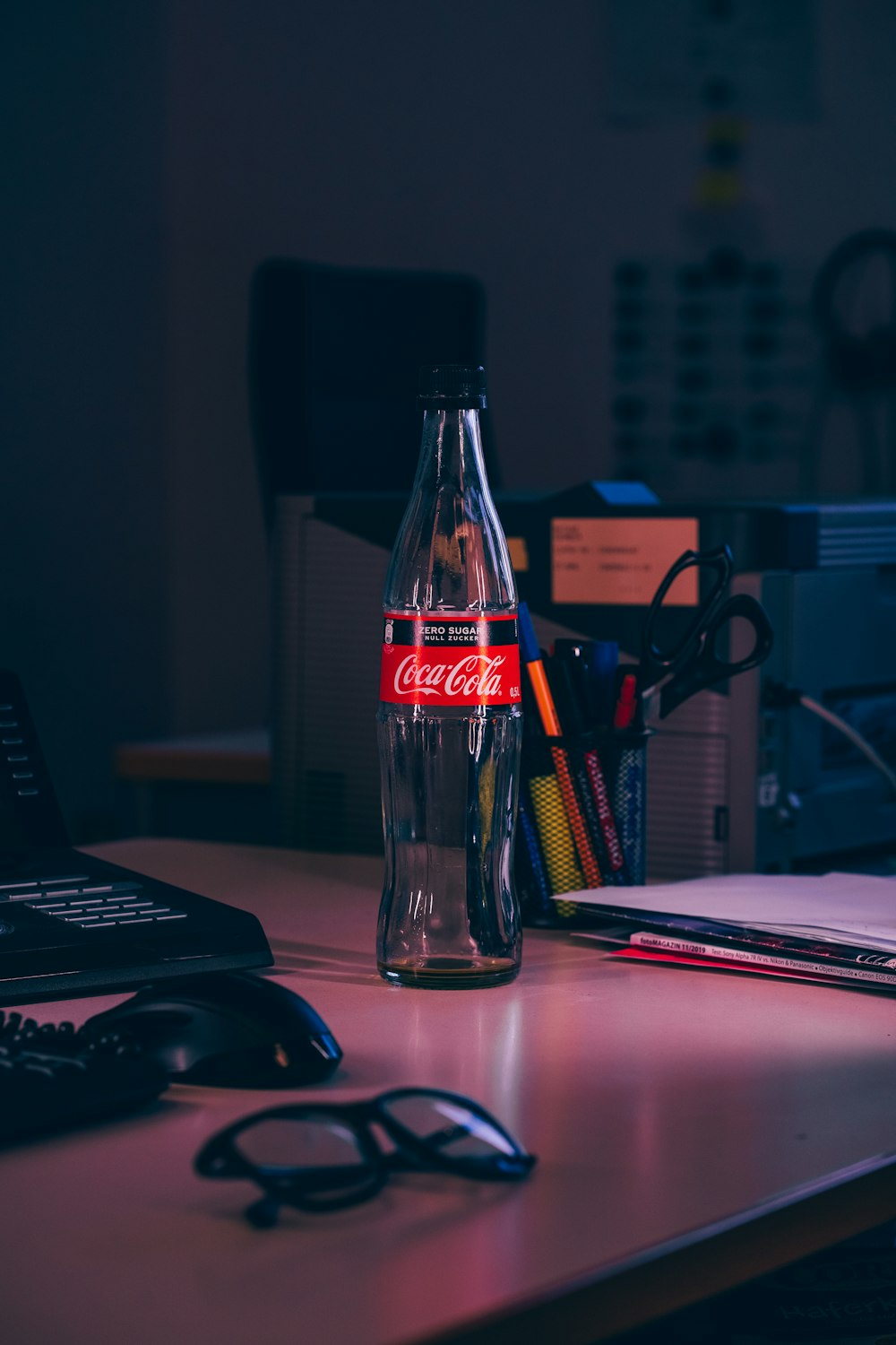 leere Coca-Cola-Flasche in der Nähe von Brille, Druckerpapier, Tastatur und Computermaus auf weißem Tisch