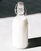 A glass bottle filled with milk is standing against a contrasting background. The bottle has a metal clasp and a rubber stopper at the top.