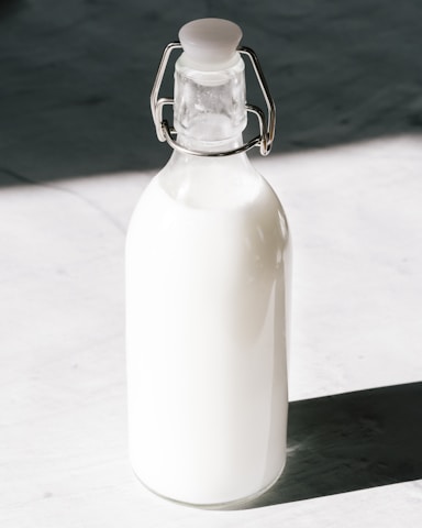 A glass bottle filled with milk is standing against a contrasting background. The bottle has a metal clasp and a rubber stopper at the top.