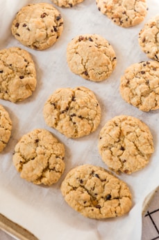 Freshly baked cookies with a golden brown color rest on a baking sheet lined with parchment paper. The cookies have a cracked surface and are studded with chocolate chips.