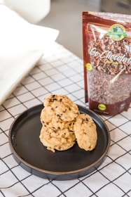 A black plate holds a stack of chocolate chip cookies placed on a white and black grid-patterned tablecloth. Next to the plate is an upright package of organic chocolate labeled 'Go Chocolaty.' The background features a slightly blurred area with neutral colors.