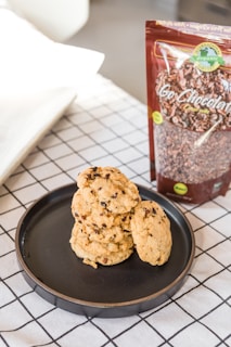 A black plate holds a stack of chocolate chip cookies placed on a white and black grid-patterned tablecloth. Next to the plate is an upright package of organic chocolate labeled 'Go Chocolaty.' The background features a slightly blurred area with neutral colors.
