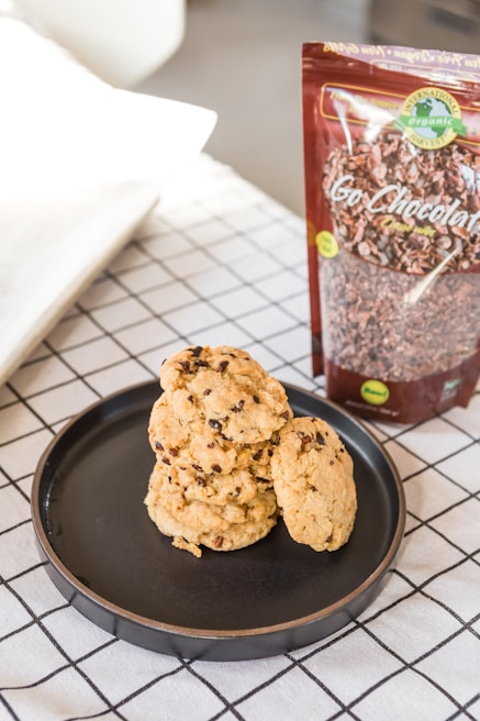 A black plate holds a stack of chocolate chip cookies placed on a white and black grid-patterned tablecloth. Next to the plate is an upright package of organic chocolate labeled 'Go Chocolaty.' The background features a slightly blurred area with neutral colors.