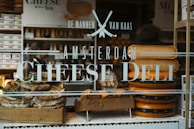 A storefront window features text advertising an Amsterdam cheese deli. Behind the glass, large wheels of cheese are stacked beside a wooden crate filled with bread or pastries garnished with herbs. The interior showcases numerous shelves filled with cheese and related products.