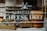 A storefront window features text advertising an Amsterdam cheese deli. Behind the glass, large wheels of cheese are stacked beside a wooden crate filled with bread or pastries garnished with herbs. The interior showcases numerous shelves filled with cheese and related products.