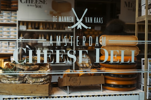 A storefront window features text advertising an Amsterdam cheese deli. Behind the glass, large wheels of cheese are stacked beside a wooden crate filled with bread or pastries garnished with herbs. The interior showcases numerous shelves filled with cheese and related products.