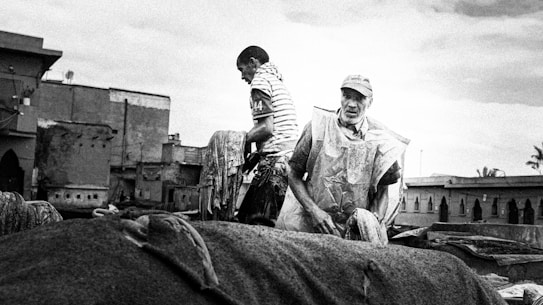 Two men are working, presumably in a traditional tanner's yard. One man is wearing a striped shirt and the other is in a cap and protective clothing. The scene is set against a backdrop of old, rustic buildings, suggesting an industrial or manual labor setting.