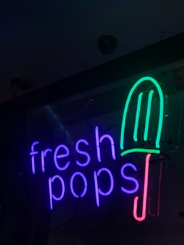 Neon sign featuring the words 'fresh pops' with a popsicle graphic. The text is glowing in purple, and the popsicle is depicted with a green top and a red handle.