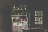 A rustic kitchen with a wooden cabinet filled with ceramic pots and glass jars. The cabinet has a weathered look, adding to the vintage atmosphere. A small window lets in some natural light, revealing greenery outside.