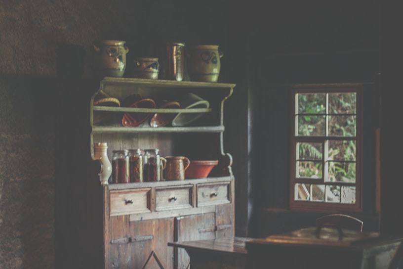 A cozy farmhouse kitchen corner featuring a rustic wooden hutch cabinet filled with enamel canisters and woven jute mats.
