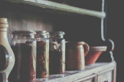 A rustic wooden shelf holds several glass jars filled with various dried goods. There is also a ceramic jug and a bowl, contributing to an overall vintage aesthetic. The lighting is soft and creates a warm, nostalgic feel.