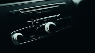 A close-up of a car's multimedia control panel, featuring several buttons and a prominent volume knob. The branding above the buttons indicates it's from Audi, with options labeled for media, car, and radio features.
