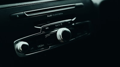 A close-up of a car's multimedia control panel, featuring several buttons and a prominent volume knob. The branding above the buttons indicates it's from Audi, with options labeled for media, car, and radio features.