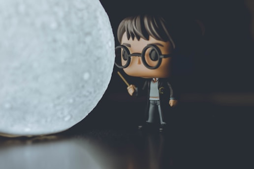 A small, stylized figurine resembling a character in wizard attire stands on a dark surface, holding a wand. The figurine has circular glasses and short hair. In the foreground, a spherical object, resembling a textured moon or a glowing orb, emits a soft light.